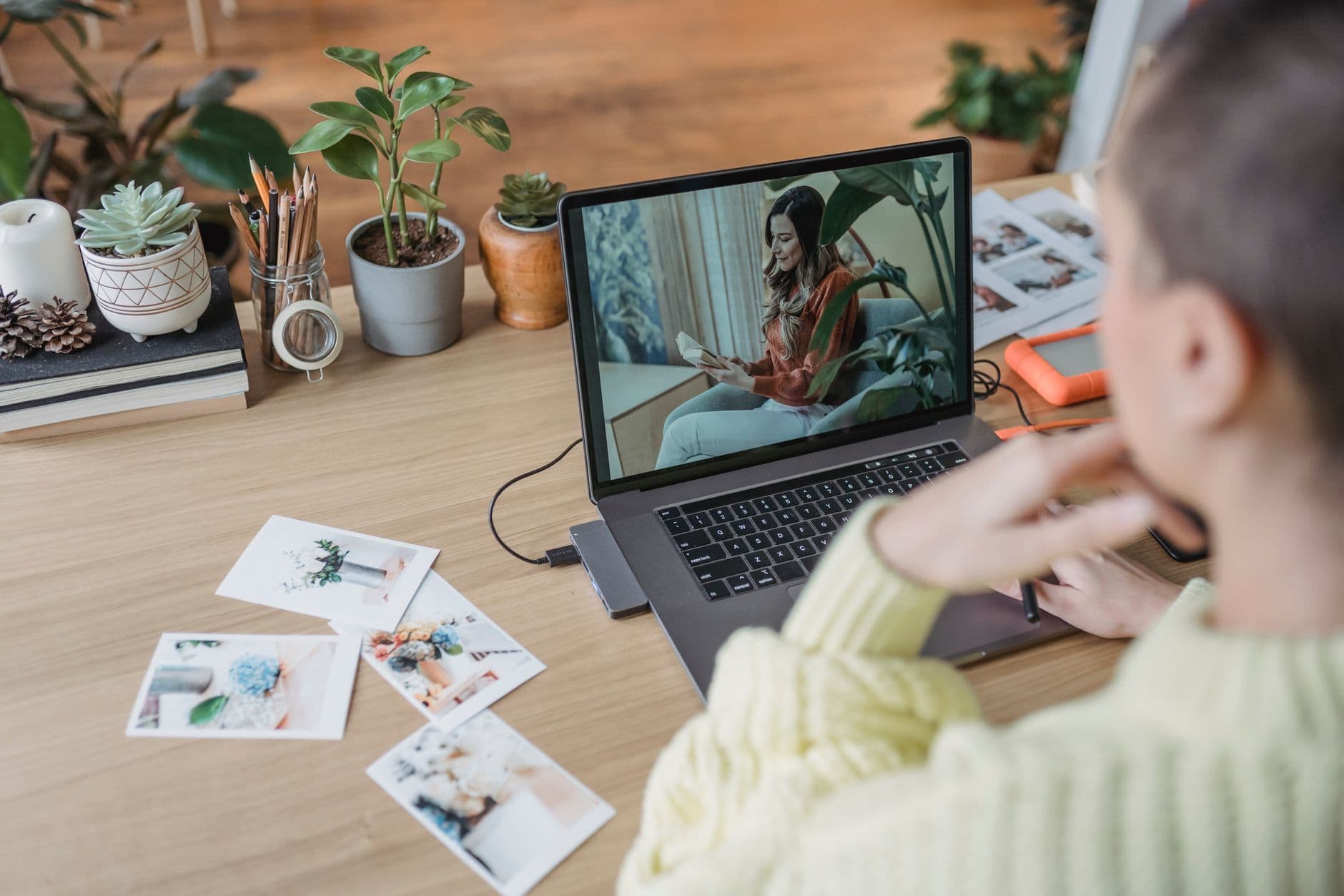 Crop unrecognizable female photographer retouching photo on netbook while sitting at wooden table with printed images and small potted plants