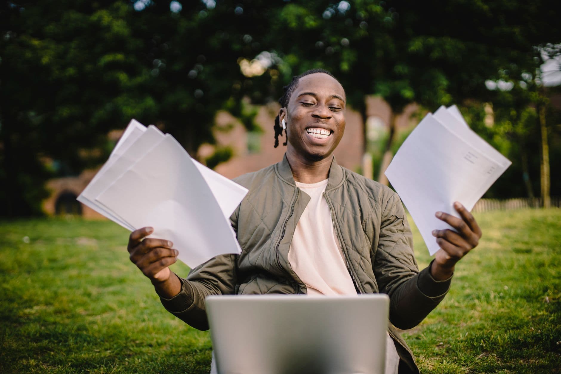 Super positive black student sitting in green park with application papers and laptop and smiling happily after learning news about being accepted to university