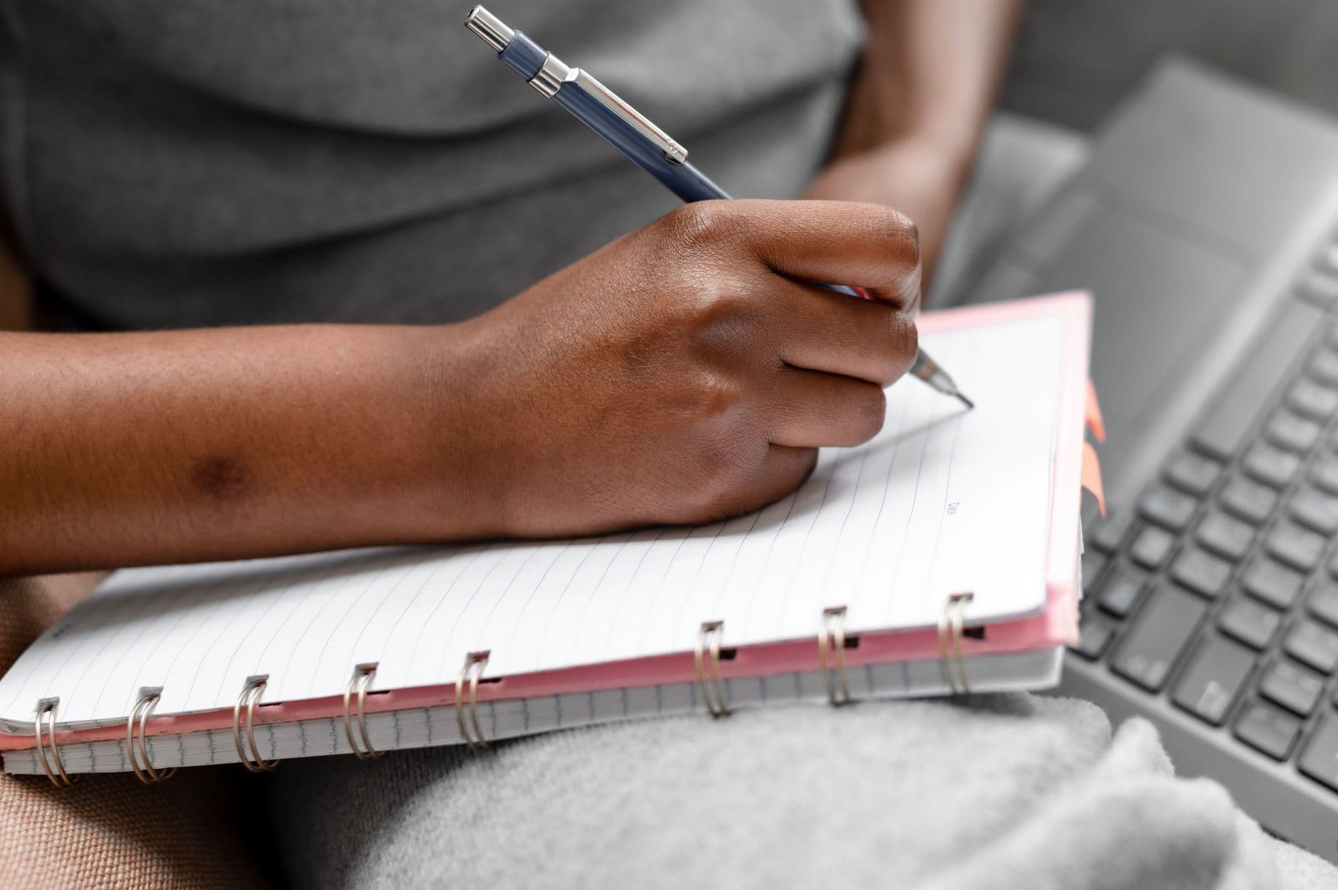 A person writing in a spiral notebook beside a laptop, focusing on note-taking and study.
