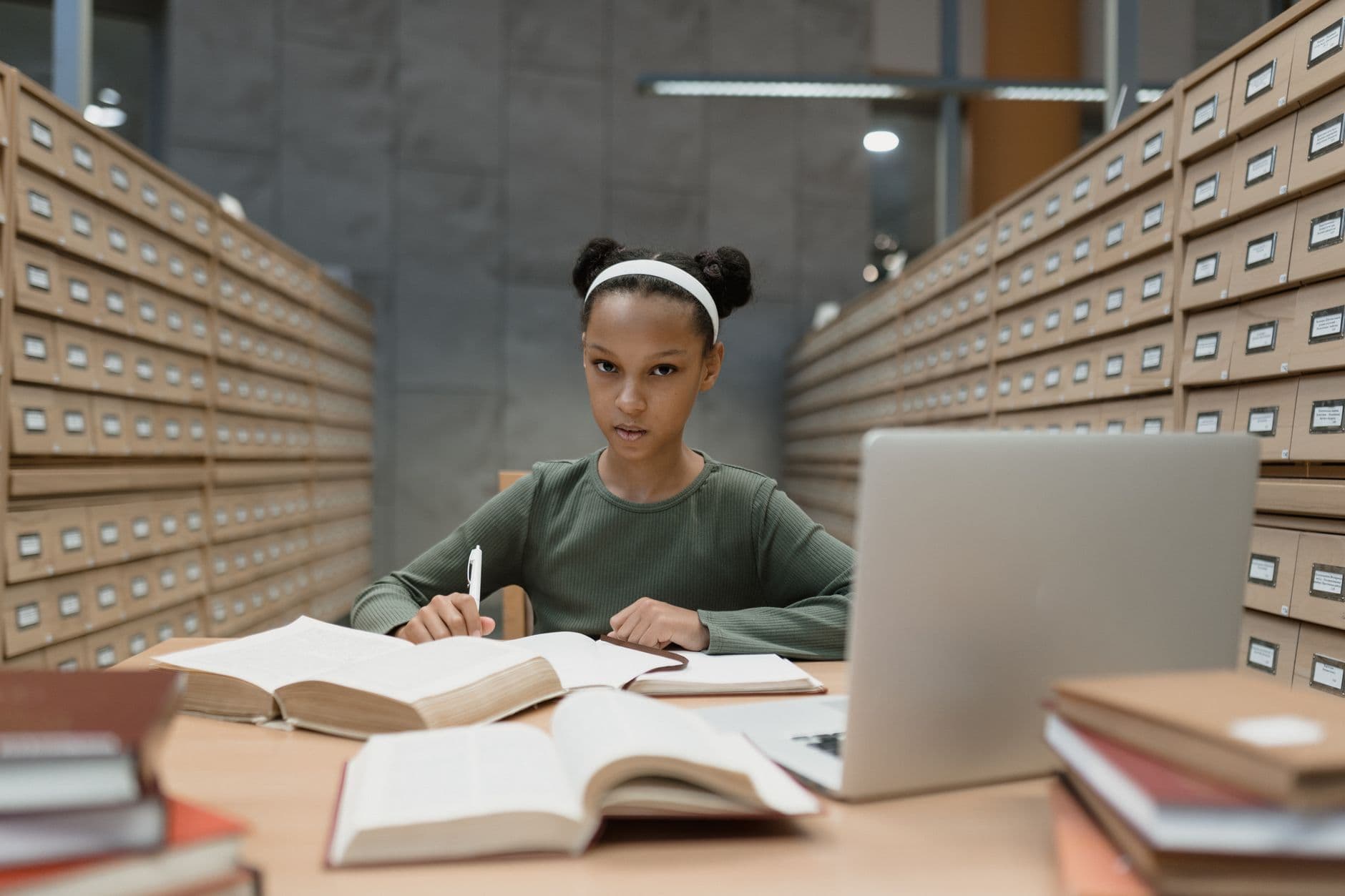 A focused young girl studying at a library desk with books and a laptop.