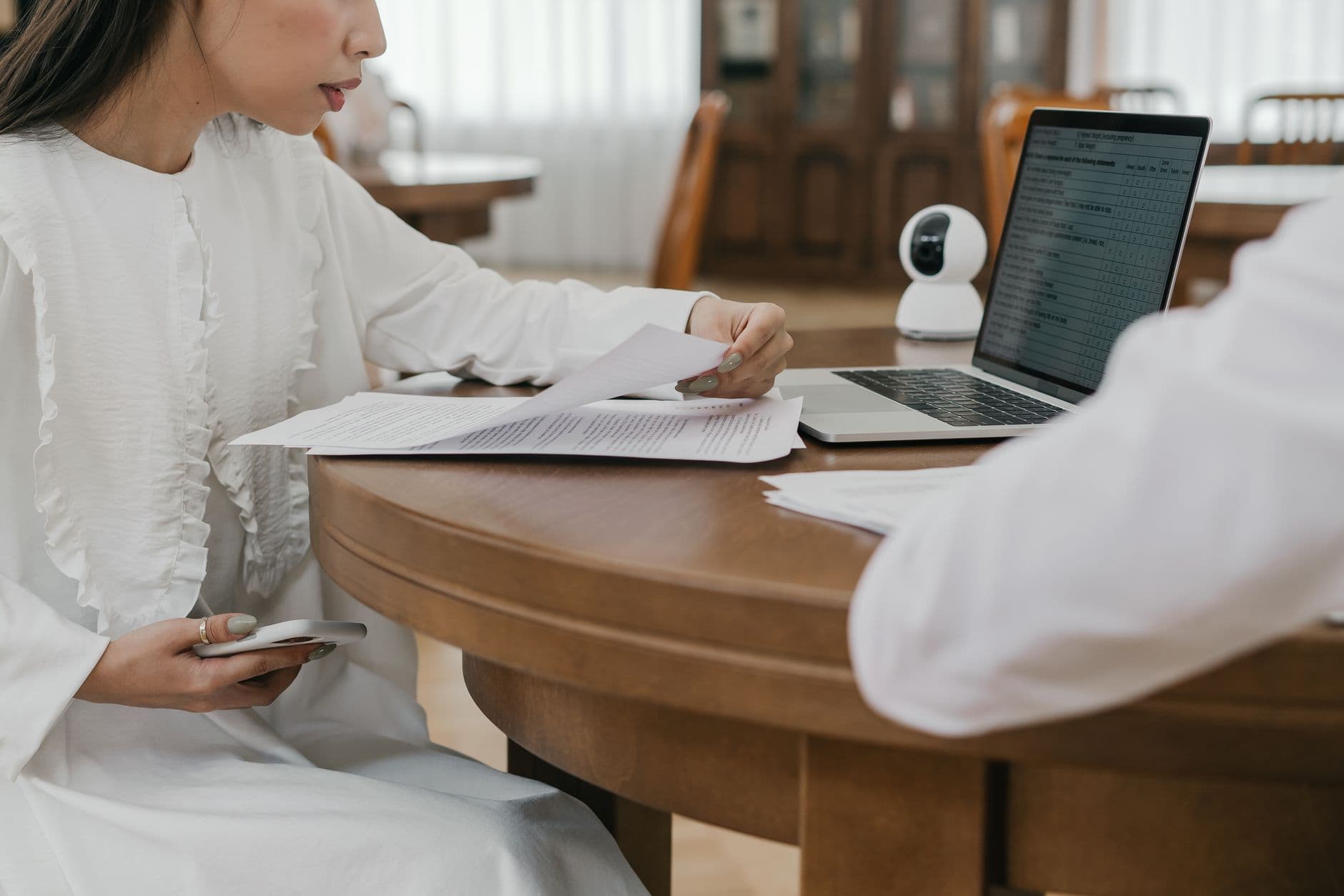 A woman in white reviews documents next to a laptop on a wooden table indoors.