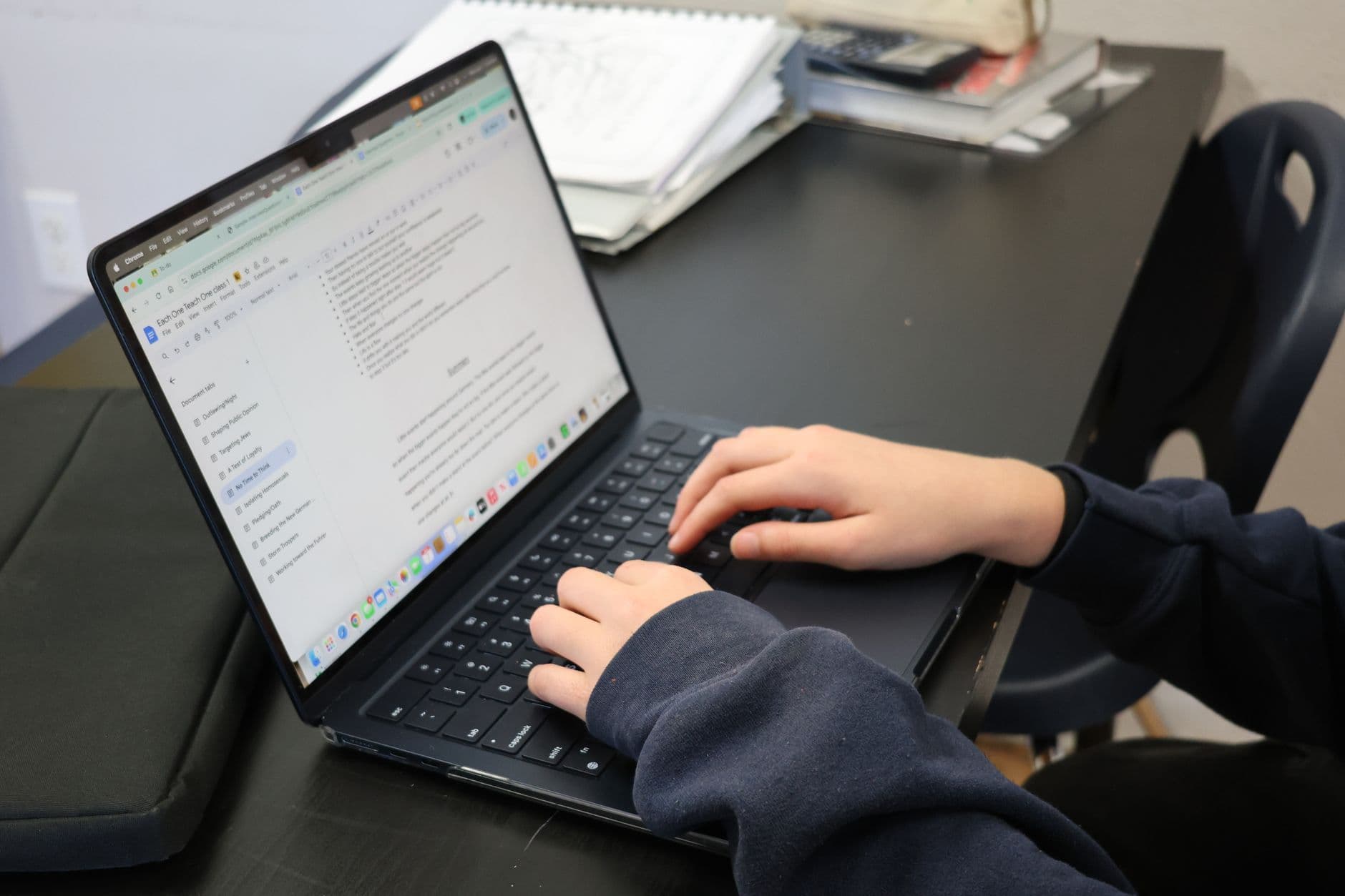 Person typing on a laptop at a desk with papers and books nearby.