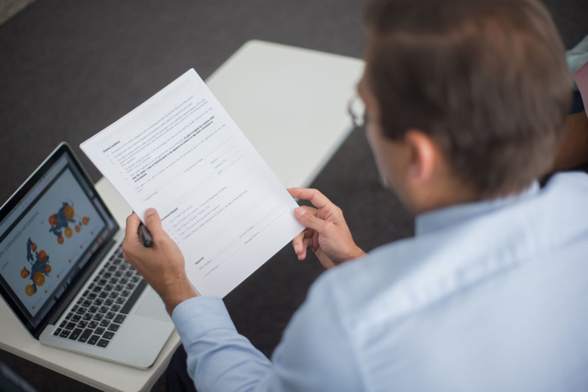 Professional in blue shirt reviewing documents next to laptop displaying charts.