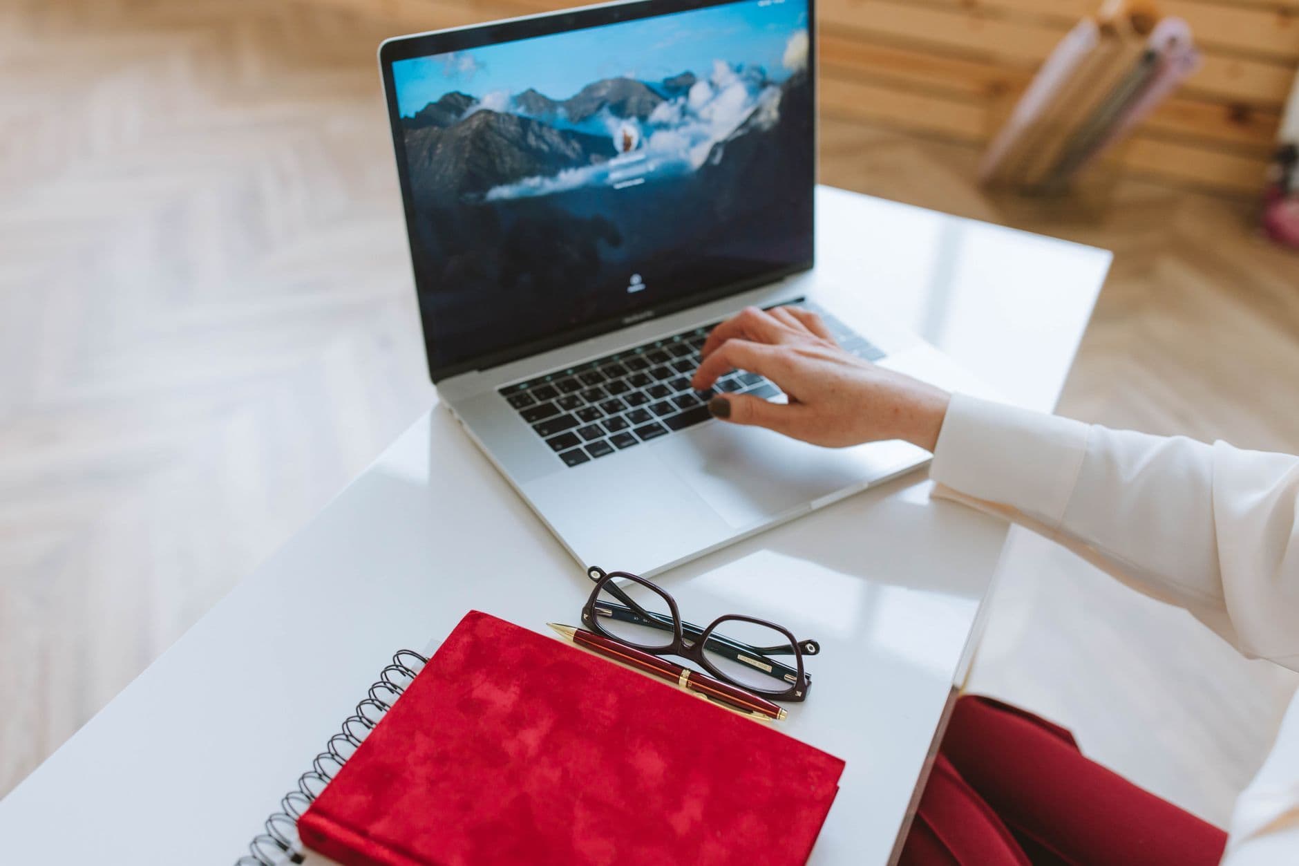 A person working on a laptop with a red notebook and glasses on a white table.