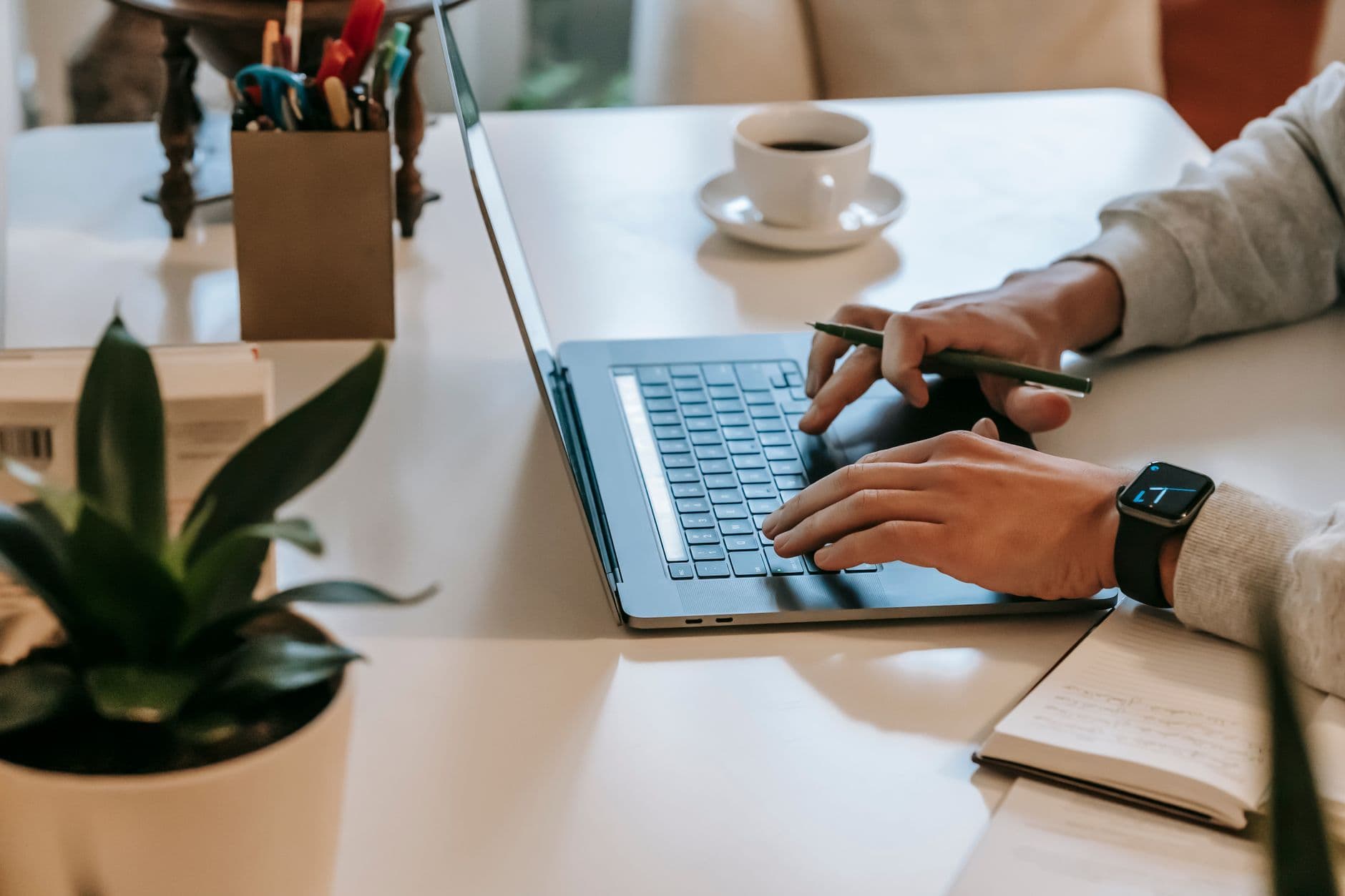 High angle side view of crop unrecognizable guy with smartwatch working remotely on laptop at table near cup of coffee on saucer near green potted plant and notebook in room