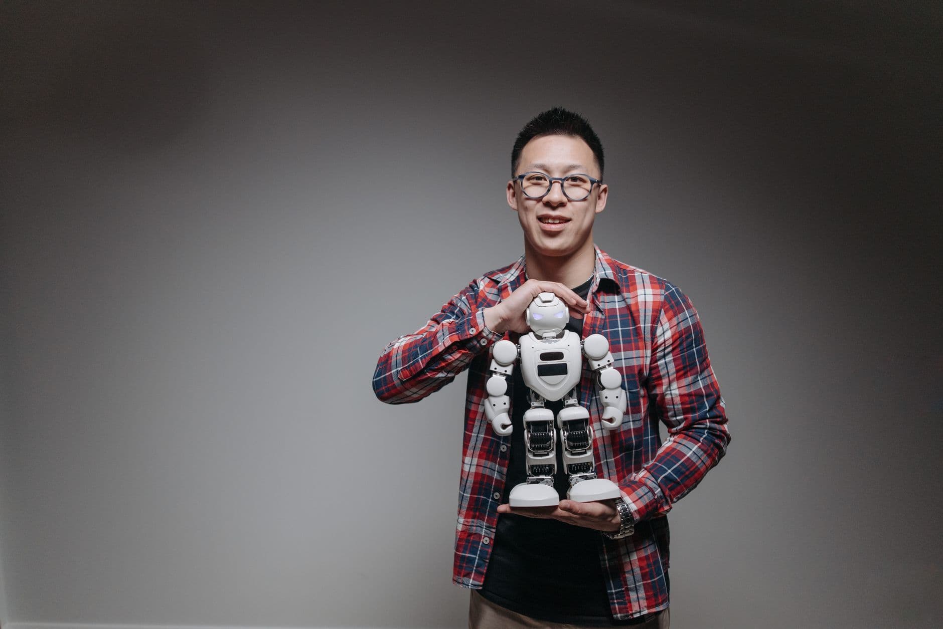 Asian man with eyeglasses holding a toy robot in a studio with a gray background.