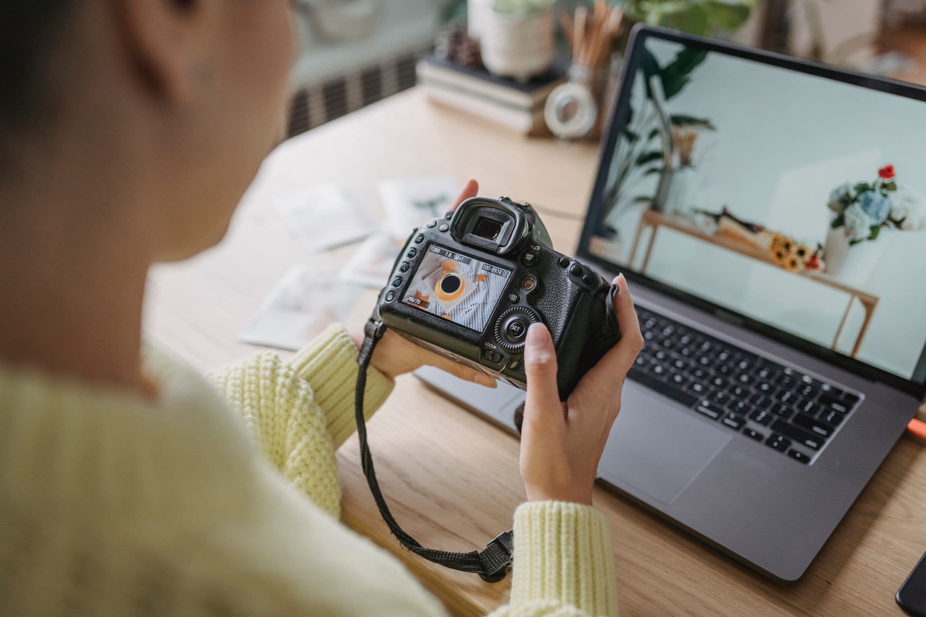 Unrecognizable crop female photographer looking at screen of photo camera while sitting at wooden table with netbook during work in light room